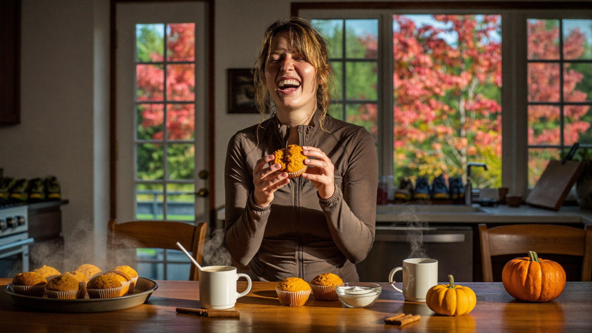A smiling runner in an autumn running jacket breaks open a steaming pumpkin muffin at a sunlit kitchen counter, with a tray of muffins, a frothy coffee and a small pumpkin beside her and fall trees outside the window.